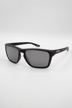 A pair of sleek, black Oakley Sylas Polarized sunglasses featuring a rectangular frame and dark grey lenses. The right temple showcases a small white "O" logo. Enhanced with Prizm lens technology, these sunglasses are positioned at an angle on a plain white background.
