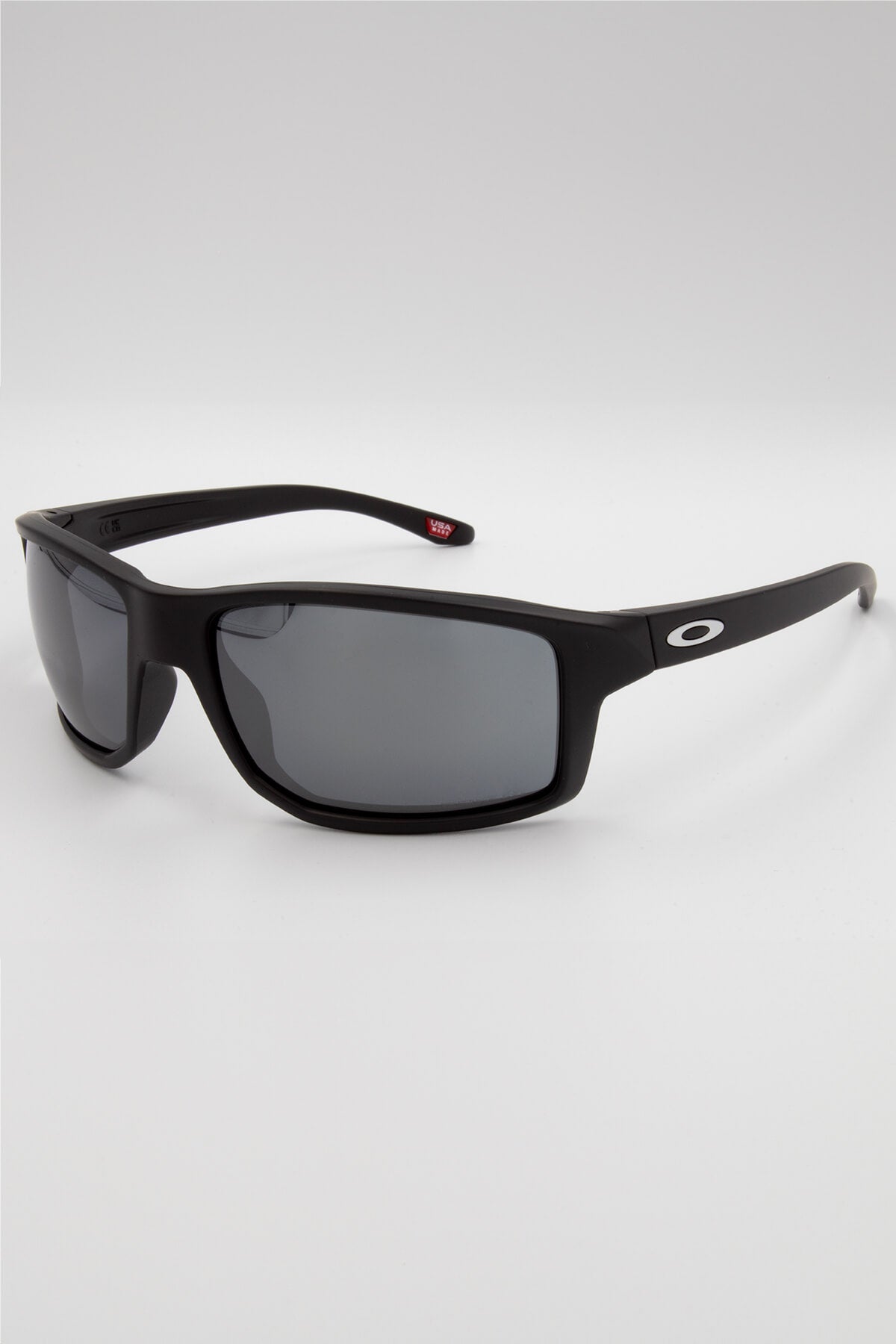 The Oakley Gibston features a black rectangular frame made from lightweight O Matter, dark lenses, and a thick matte finish. A white Oakley logo accents the temple arm. The sunglasses are shown on a plain, light background.