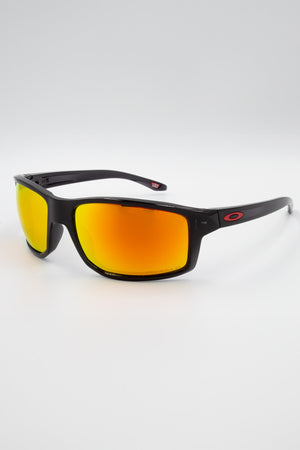 A pair of Oakley Gibston Polarized sunglasses, featuring black frames with orange-red mirrored Prizm lenses and red “O” logos on the temples, shown against a white background.