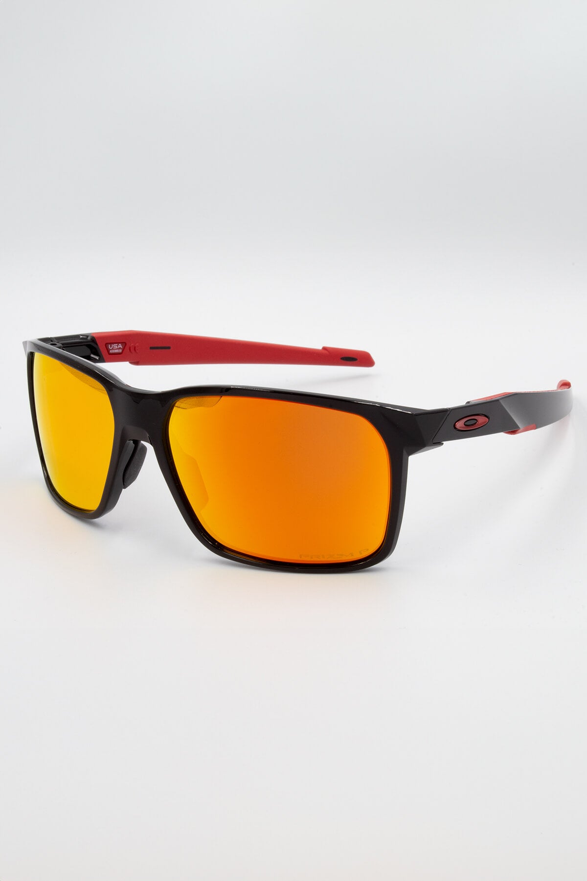 The Oakley Portal X Polarized sunglasses feature a sleek black frame with Ruby Polarized lenses and red temples showcasing the Oakley logo. Enhanced with Prizm lens technology for better contrast and clarity, they are stylishly angled on a white surface.