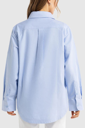 A person is seen from behind wearing the oversized ORTC Adelaide Oxford Shirt in light blue, made from premium cotton, with their hair partially visible and white pants showing beneath the shirt hem.