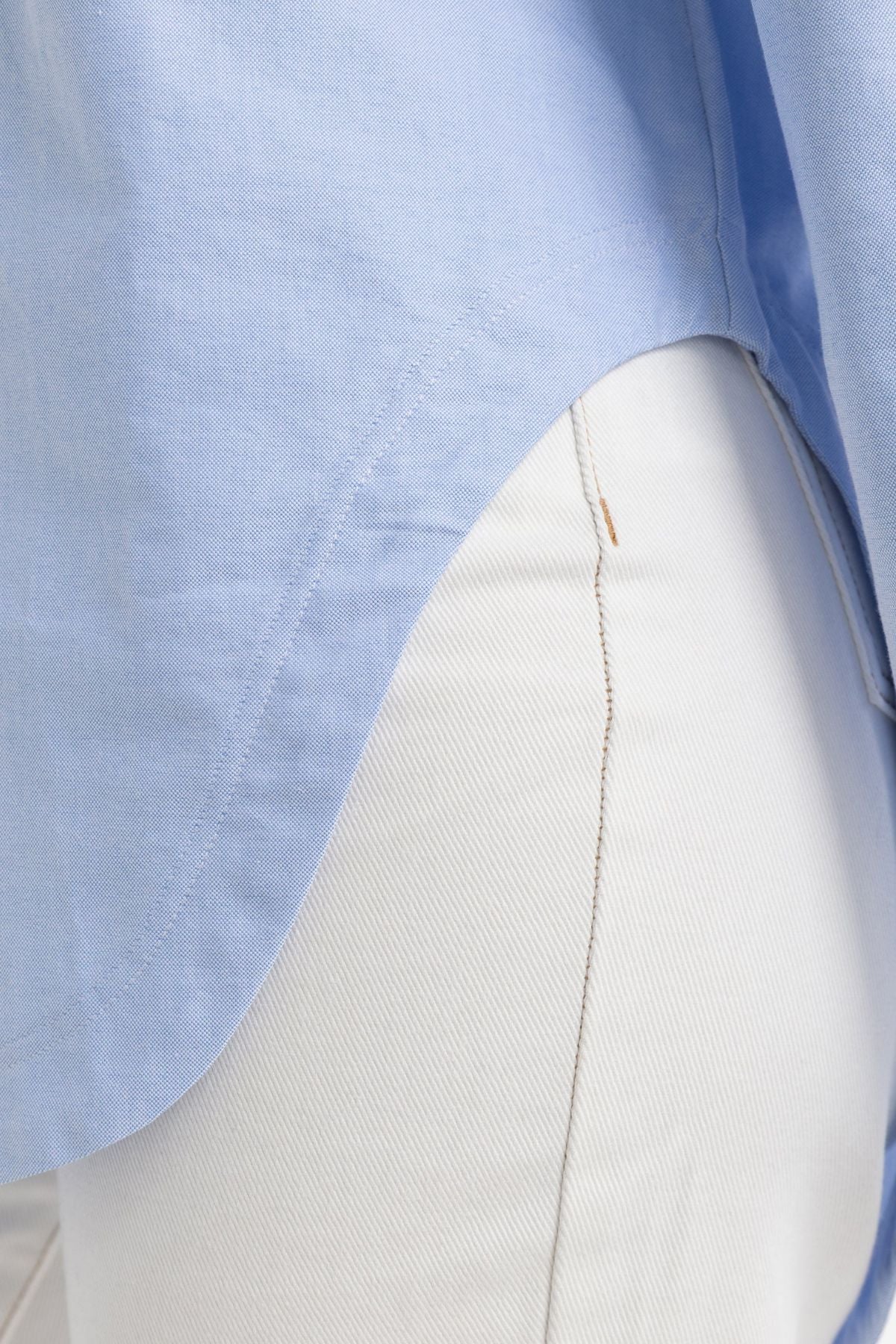 A close-up of a person wearing the ORTC Adelaide Oxford Shirt in light blue, styled untucked over white pants, highlighting the shirt’s curved hem and high-quality cotton fabric alongside the seam detail of the pants.