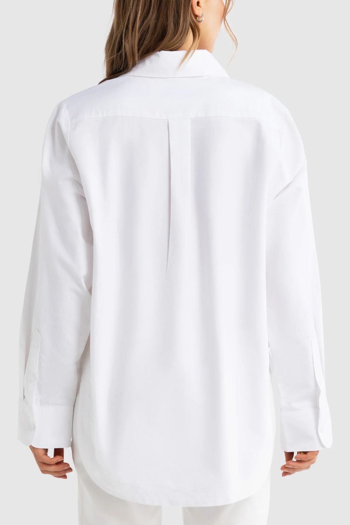 A person with long hair is seen from behind, dressed in the ORTC Adelaide Oxford Shirt—an oversized, premium cotton long-sleeve shirt—and white pants, set against a plain light gray background.