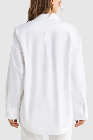 A person with long hair is seen from behind, dressed in the ORTC Adelaide Oxford Shirt—an oversized, premium cotton long-sleeve shirt—and white pants, set against a plain light gray background.
