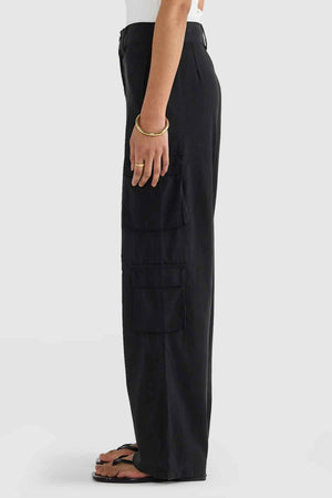 The individual is pictured in a side profile sporting the ORTC Arden Cargo Pants in black, which feature cargo pockets on both the thigh and calf. They pair this with a white top, black sandals, and a gold bracelet against a clean white background.