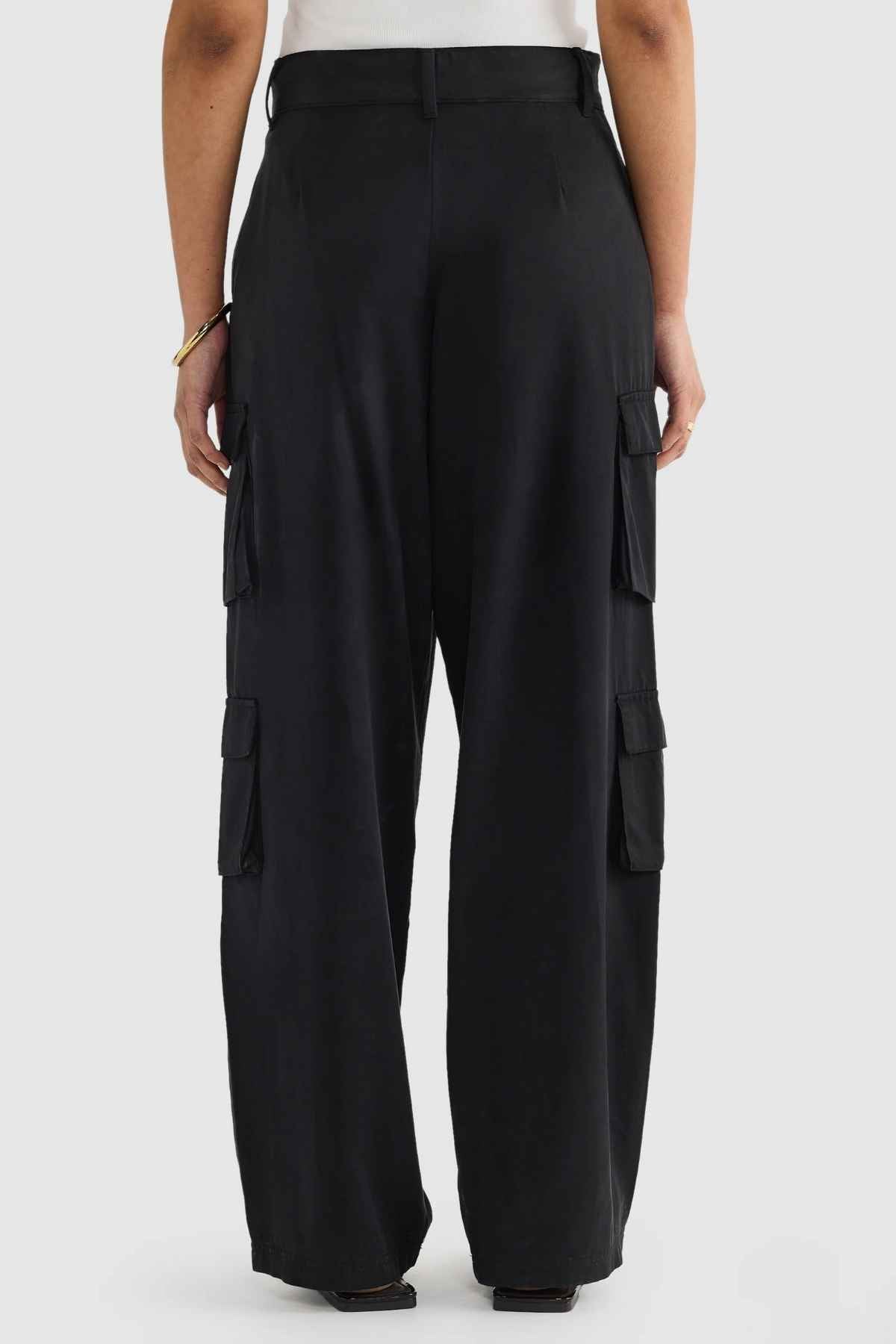 A person stands against a plain background, showcasing ORTC Arden Cargo Pants made from a blend of Tencel and cotton, featuring several cargo pockets. They've combined these pants with a white shirt and accessorized with a bracelet on their left wrist.