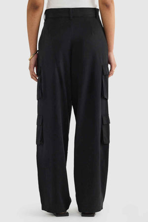 A person stands against a plain background, showcasing ORTC Arden Cargo Pants made from a blend of Tencel and cotton, featuring several cargo pockets. They've combined these pants with a white shirt and accessorized with a bracelet on their left wrist.