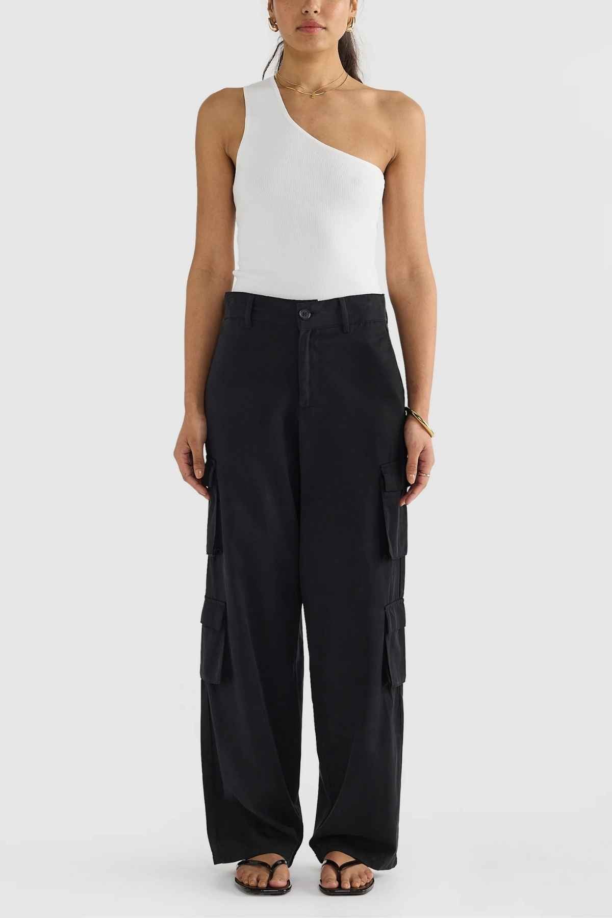 A person dressed in a white one-shoulder top, black ORTC Arden Cargo Pants featuring cargo pockets, and black sandals against a plain white background.