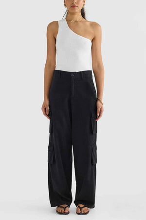 A person dressed in a white one-shoulder top, black ORTC Arden Cargo Pants featuring cargo pockets, and black sandals against a plain white background.