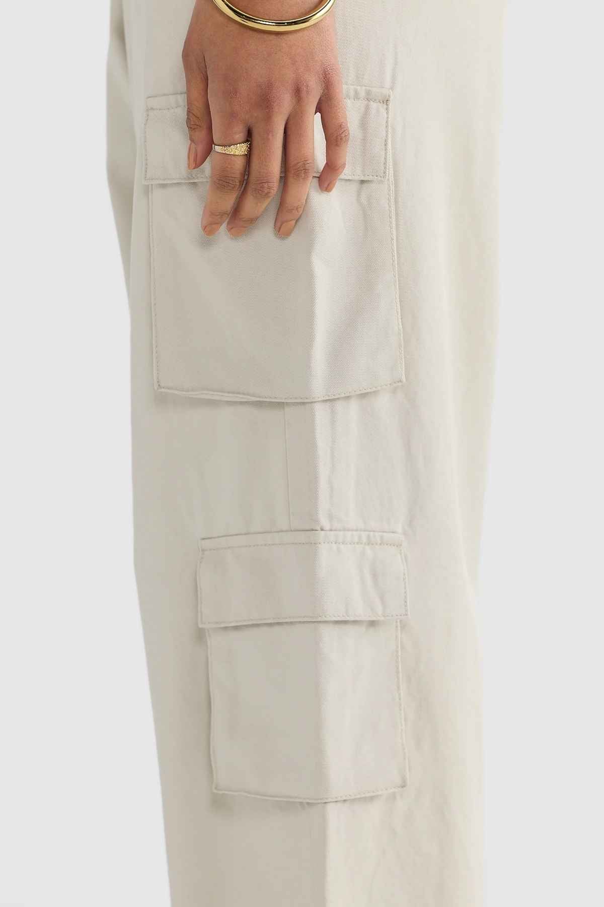 The close-up highlights an individual wearing ORTC Arden Cargo Pants in cream, crafted from tencel-cotton fabric. The shot neatly emphasizes two flap cargo pockets as the person places their hand on one, showcasing a bracelet and ring against a sleek light gray background.