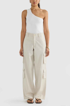 A person stands against a neutral background wearing the ORTC Arden Cargo Pants, crafted from a soft tencel-cotton blend in light beige. These stylish pants feature multiple cargo pockets and are paired with a chic white one-shoulder top and black sandals for a practical yet fashionable look.