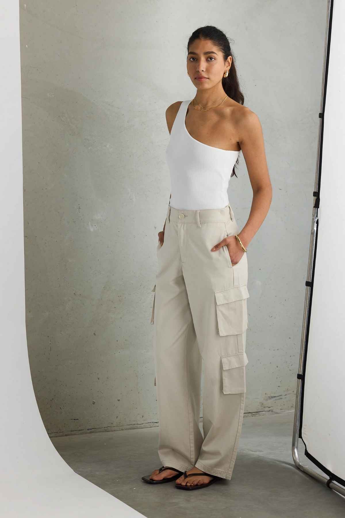 A person is positioned against a simple backdrop, donning a white one-shoulder top paired with the ORTC Arden Cargo Pants, made from tencel-cotton material. The beige cargo pants come with utility pockets and are paired with black sandals and gold jewelry. With long dark hair, they pose with one hand casually tucked into a pocket.