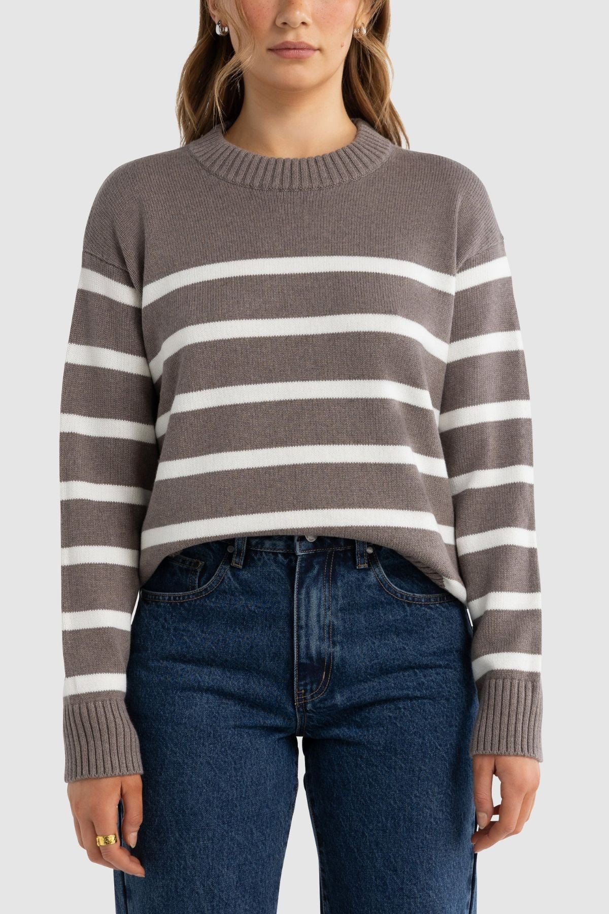 A person wears the ORTC Astrid Crew Knit—a taupe and white striped cotton cashmere sweater—and blue jeans, standing facing forward against a plain light gray background.