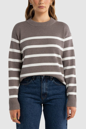 A person wears the ORTC Astrid Crew Knit—a taupe and white striped cotton cashmere sweater—and blue jeans, standing facing forward against a plain light gray background.