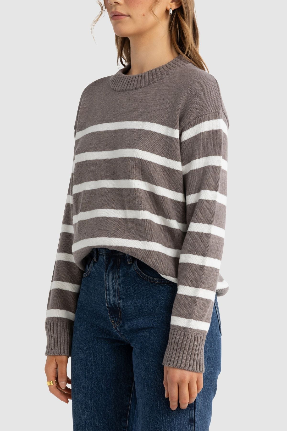 A woman wears the ORTC Astrid Crew Knit, a relaxed fit sweater with white horizontal stripes, paired with blue jeans and standing against a plain light gray background.