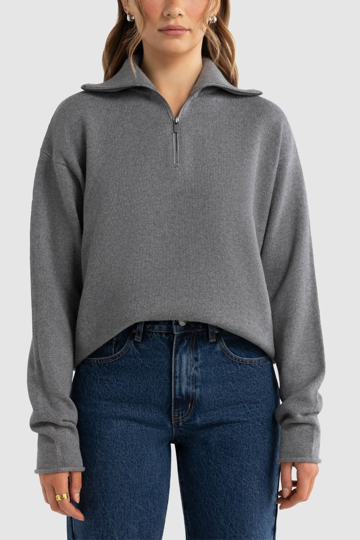 Someone stands against a light gray background wearing the ORTC Astrid Knit Quarter Zip, a gray cotton-cashmere blend sweater with a folded collar, relaxed fit, and long sleeves, paired with high-waisted blue jeans. Ideal for layering.
