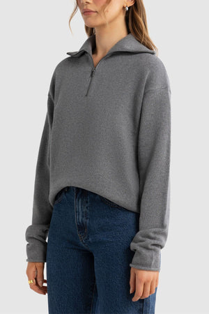 A woman wears the ORTC Astrid Knit Quarter Zip sweater in gray, featuring a collar and long sleeves, paired with high-waisted blue jeans against a plain light background.