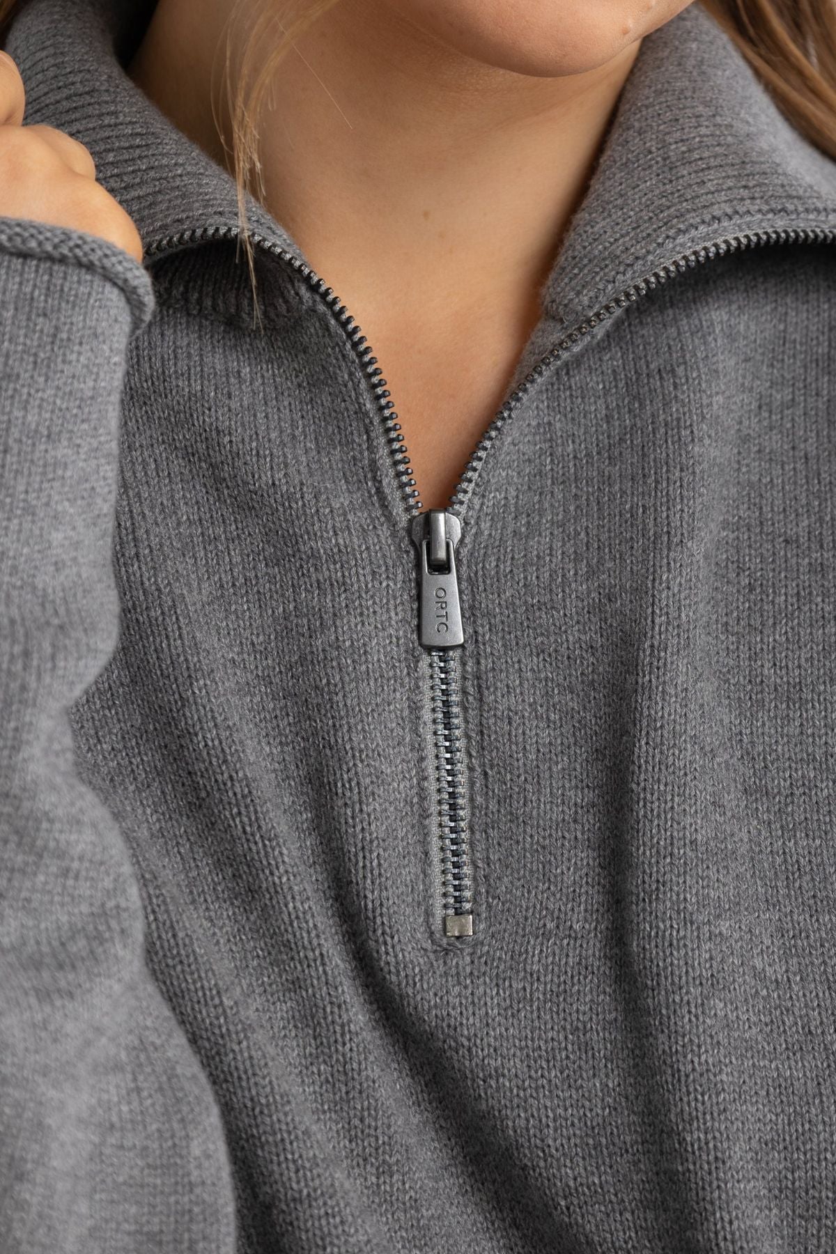 Close-up of a person wearing the ORTC Astrid Knit Quarter Zip, a grey ribbed sweater with a cotton-cashmere blend and quarter zip collar, partially unzipped to reveal part of the lower face and neck.