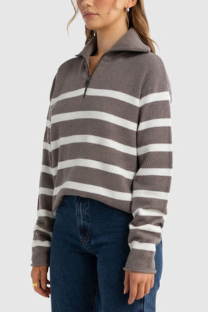 A woman wears the ORTC Astrid Knit Quarter Zip, a gray and white striped pullover, paired with dark blue jeans, standing against a plain light background.