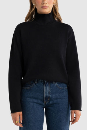 A woman stands facing forward against a light gray background, wearing the ORTC Astrid Knit Sweater in black and high-waisted blue jeans. Her hands are relaxed at her sides.