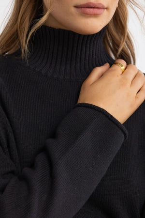 A person wearing the ORTC Astrid Knit Sweater in black touches their neck, revealing a gold ring. Only their lower face and upper torso are visible.