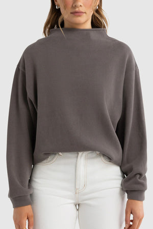 A woman stands facing forward against a plain background, wearing the ORTC Ava High Neck Jumper in dark gray with long sleeves and high-waisted white jeans.