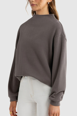 A woman wearing the ORTC Ava High Neck Jumper—a relaxed-fit, long-sleeved gray top—paired with high-waisted white jeans, standing against a plain light background.