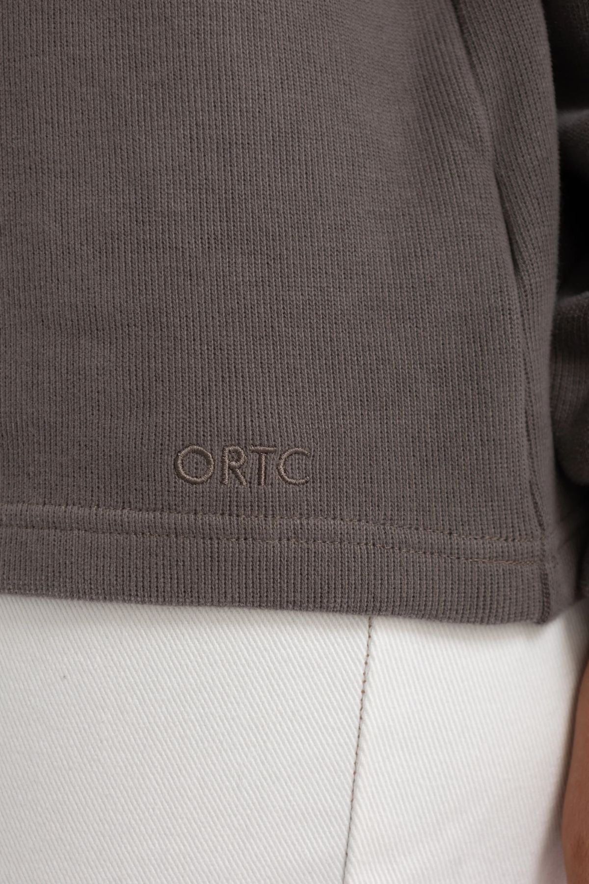 Close-up of the hem of the ORTC Ava High Neck Jumper in brown textured brushed cotton, featuring “ORTC” embroidered above light-colored pants.
