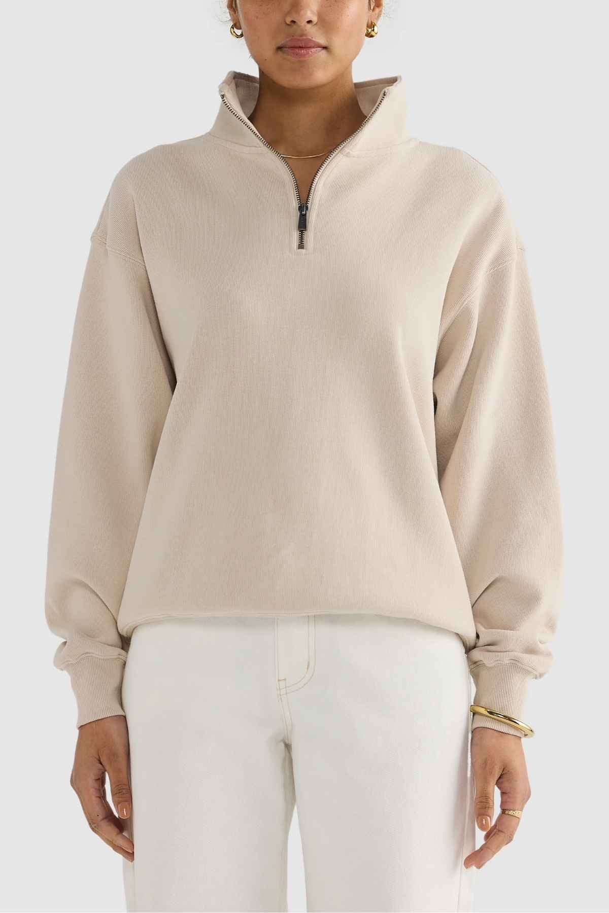 A person dressed in an ORTC Ava Ribbed Quarter Zip pullover in beige, paired with white pants, stands against a simple backdrop. This brushed cotton piece features a high collar and loose fit, making it ideal for a capsule wardrobe. The ensemble is complemented by gold earrings and a bracelet.
