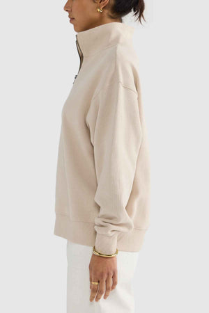 Side profile of a person wearing the ORTC Ava Ribbed Quarter Zip sweatshirt in beige, along with gold hoop earrings. They are also adorned with a gold bracelet and ring, paired stylishly with white pants. This sophisticated ensemble, perfect for any capsule wardrobe, is set against a simple light gray background.