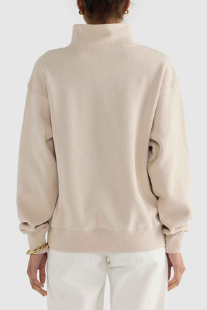 The individual is turned away from the camera, highlighting a capsule wardrobe outfit featuring the ORTC Ava Ribbed Quarter Zip in brushed cotton and white pants. The cozy long sleeves of this relaxed-fit sweater complement their hairstyle, gold hoop earrings, and bracelet against a plain gray background.