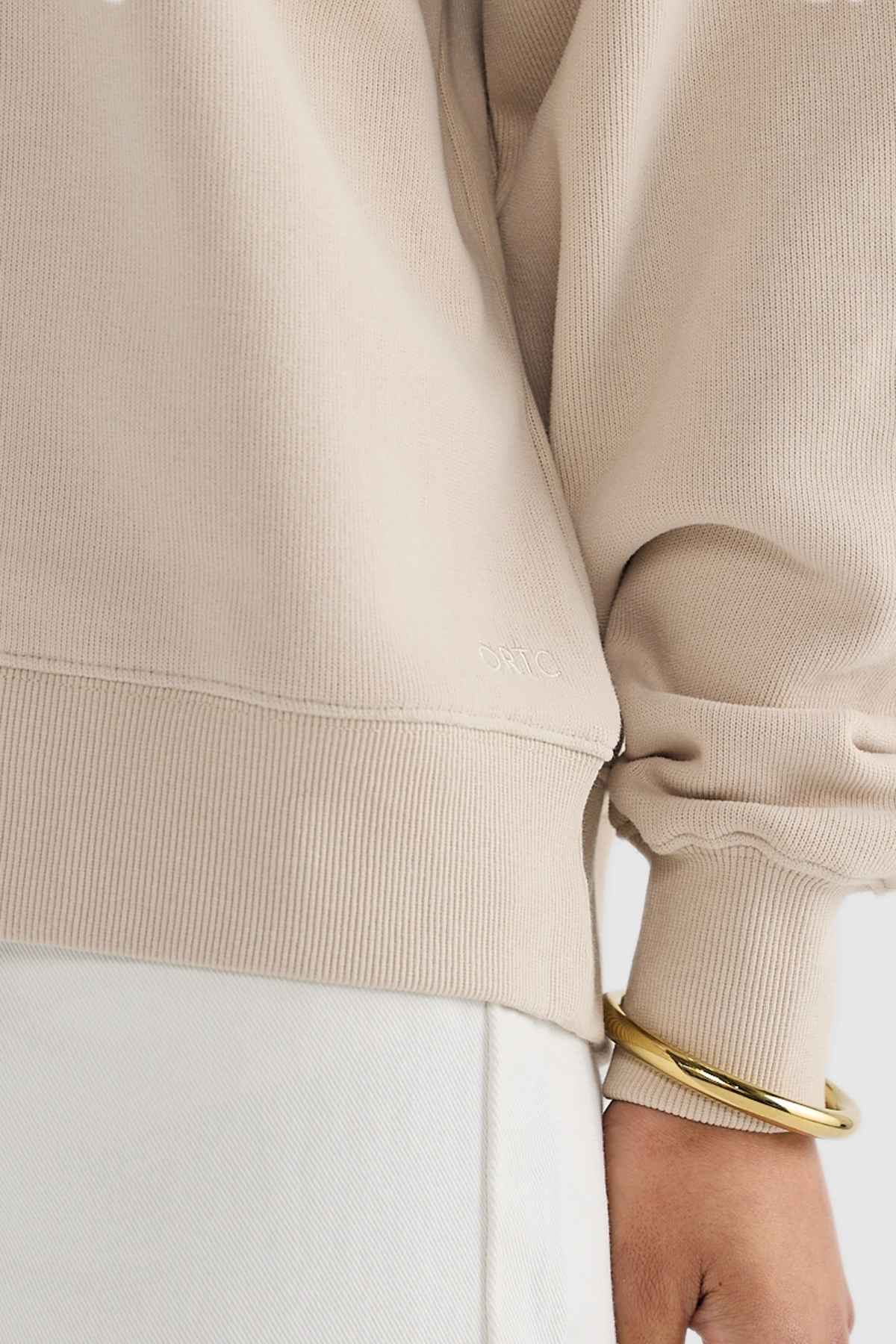 Close-up of a person wearing the ORTC Ava Ribbed Quarter Zip in beige, featuring a small embroidered logo on the back, perfect for a capsule wardrobe. With their arm by their side, they pair it with white pants and two gold bangles on their wrist, embodying effortless style in brushed cotton elegance.