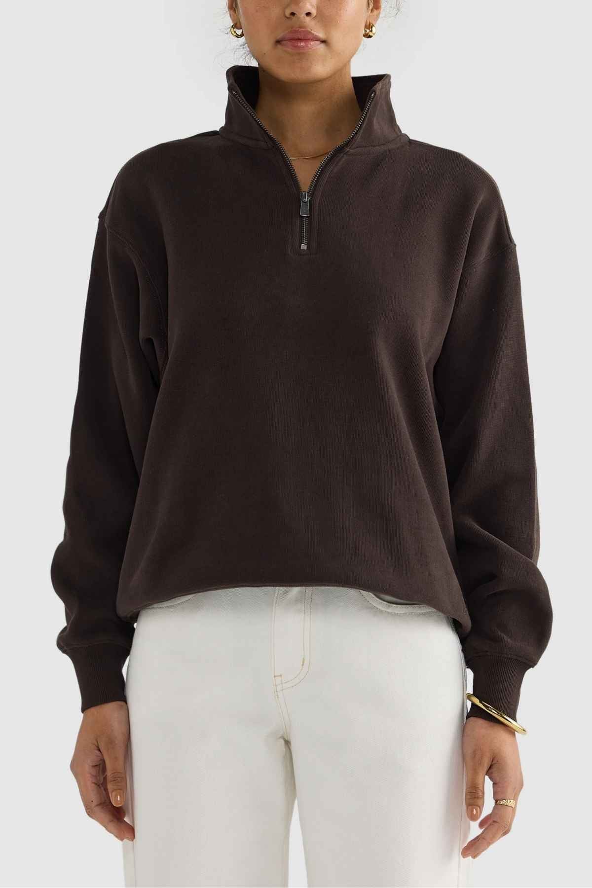 A person stands against a plain background wearing the ORTC Ava Ribbed Quarter Zip in dark brown, paired with white pants and gold hoop earrings. The pullover, crafted from brushed cotton, complements their capsule wardrobe style. They have short hair.