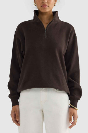 A person stands against a plain background wearing the ORTC Ava Ribbed Quarter Zip in dark brown, paired with white pants and gold hoop earrings. The pullover, crafted from brushed cotton, complements their capsule wardrobe style. They have short hair.