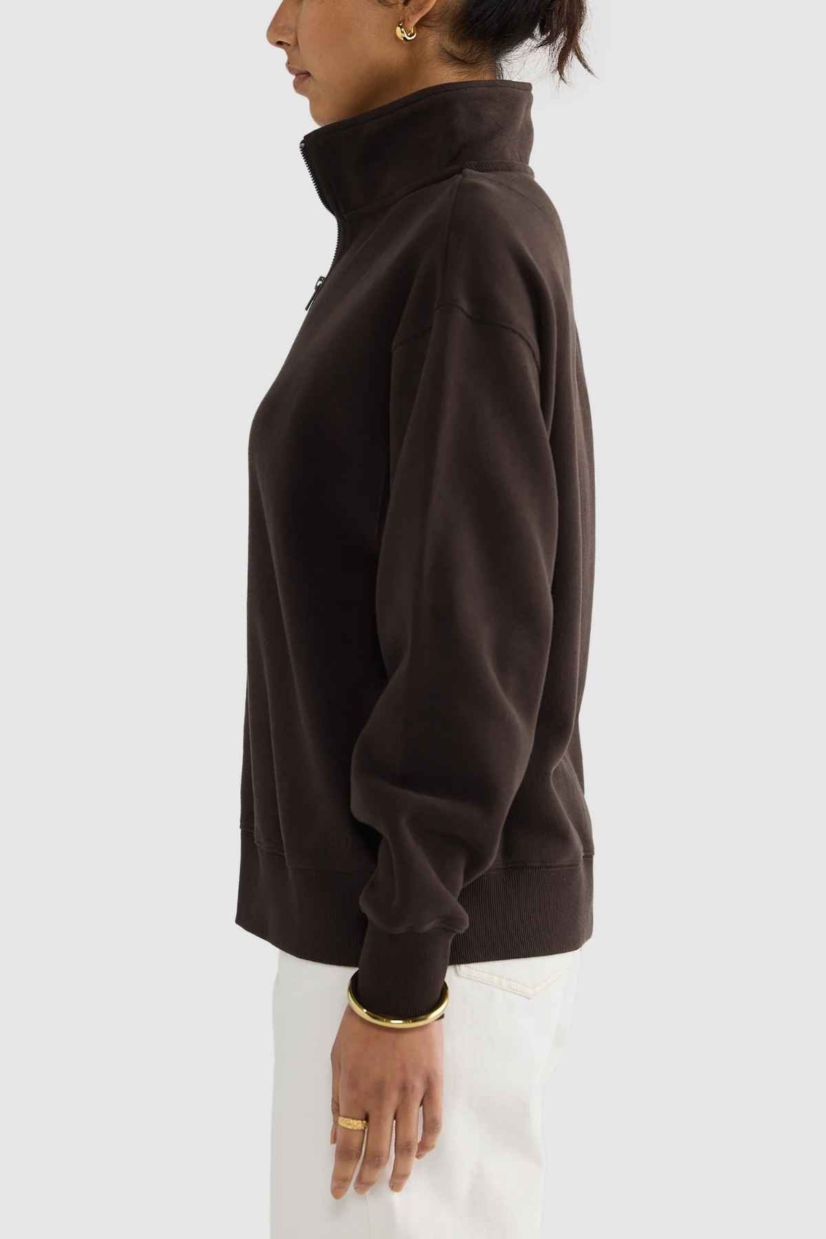 A person standing in profile against a plain background is wearing the ORTC Ava Ribbed Quarter Zip in dark brown. They have short hair and are accessorized with gold hoop earrings and a gold bracelet, creating a chic capsule wardrobe look, complemented by their white pants.