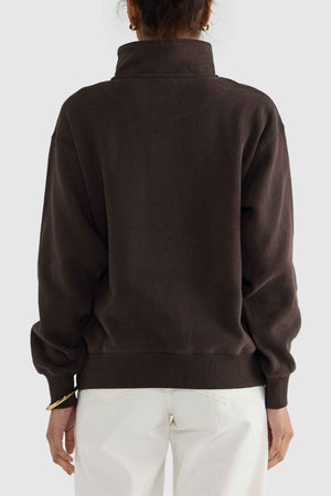 Rear view of a person wearing the ORTC Ava Ribbed Quarter Zip sweater in dark brown, paired with light-colored pants. The relaxed-fit sweater, crafted from brushed cotton, is complemented by the person's hair being tied up. The plain background accentuates the capsule wardrobe-inspired clothing.