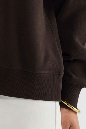 A close-up of a person's torso showcases them wearing the ORTC Ava Ribbed Quarter Zip, a dark brown brushed cotton sweatshirt with subtle text. The ensemble is completed with light-colored pants and a gold bangle on their wrist, highlighting the essence of a capsule wardrobe by emphasizing texture and color contrast.