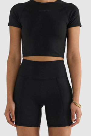 A person stands against a plain white background, wearing a black short-sleeve crop top paired with sleek high-waisted ORTC Eva 6 Inch Bike Shorts. The outfit is simple yet stylish, complemented by a gold bangle on their right wrist.