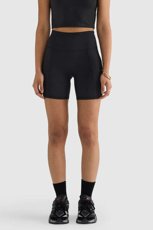 A person wearing sleek, black ORTC Eva 6 Inch Bike Shorts and a matching sleeveless top stands against a plain light gray background. Complementing the look with black socks and black-and-white sneakers, they have their arms at their sides, subtly highlighting the high-waisted design.