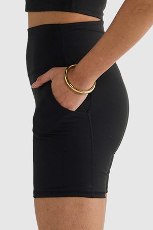A person wearing a black crop top and high-waisted ORTC Eva 6 Inch Bike Shorts with one hand in a pocket. Gold bangles adorn their wrist. The plain white background accentuates the details of the recycled polyester shorts.