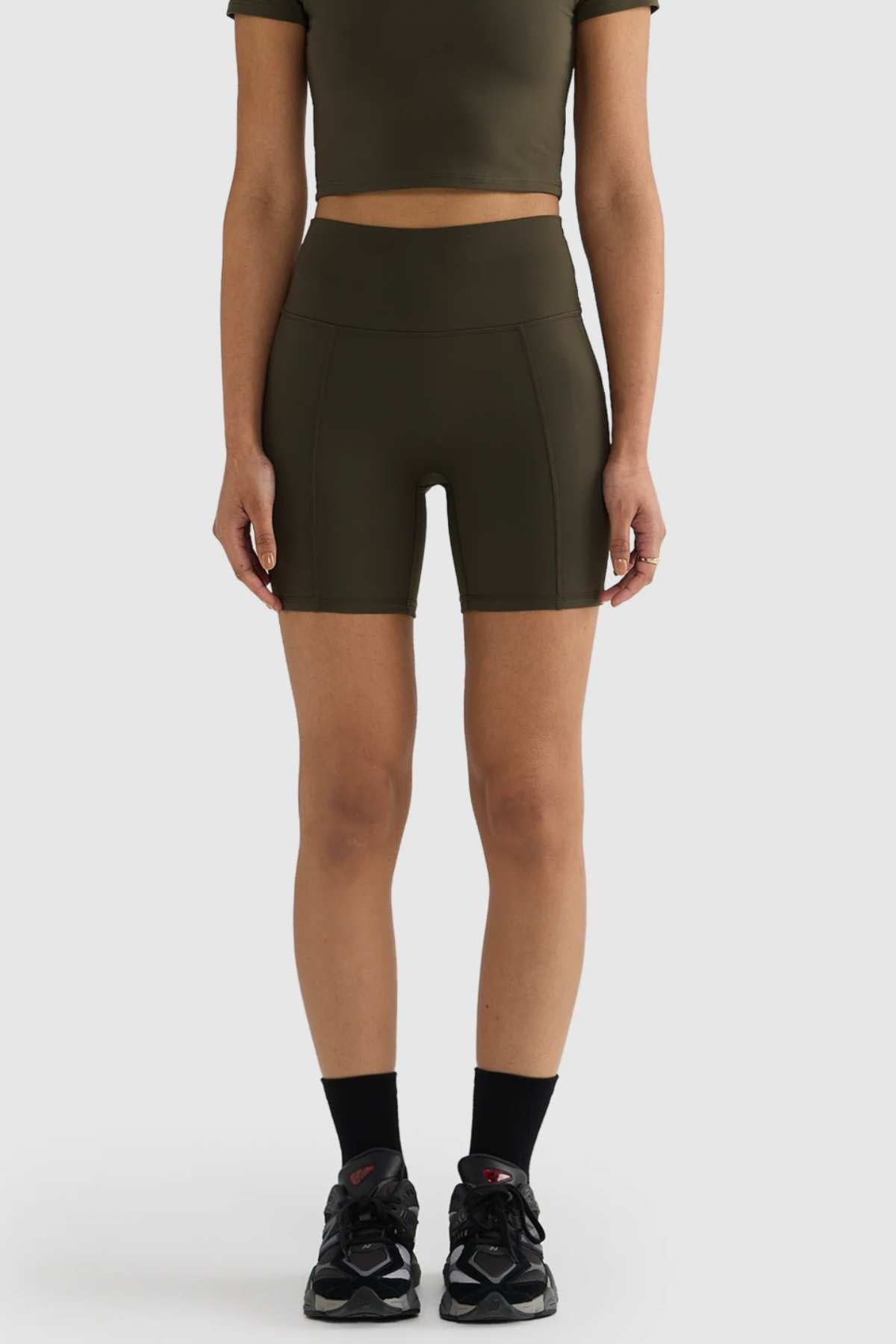 The individual is wearing a dark green crop top paired with the ORTC Eva 6 Inch Bike Shorts, featuring a comfortable high-waisted band. Complementing this look are black socks and sneakers. They are positioned against a plain light gray background.