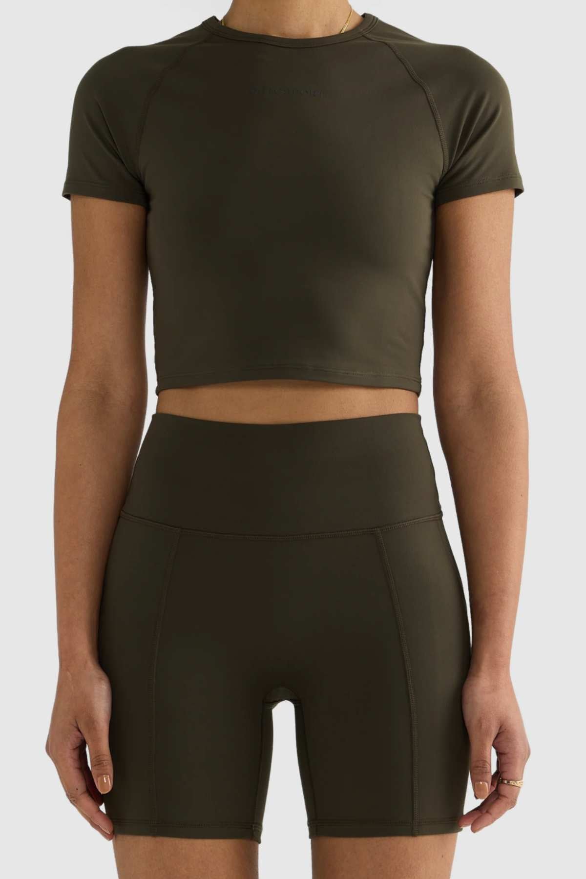 A person wearing a matching dark green fitted crop top and high-waisted ORTC Eva 6 Inch Bike Shorts. The minimal, sporty outfit features short sleeves and a smooth recycled polyester fabric texture. The background is plain and light-colored, highlighting the sleek design.