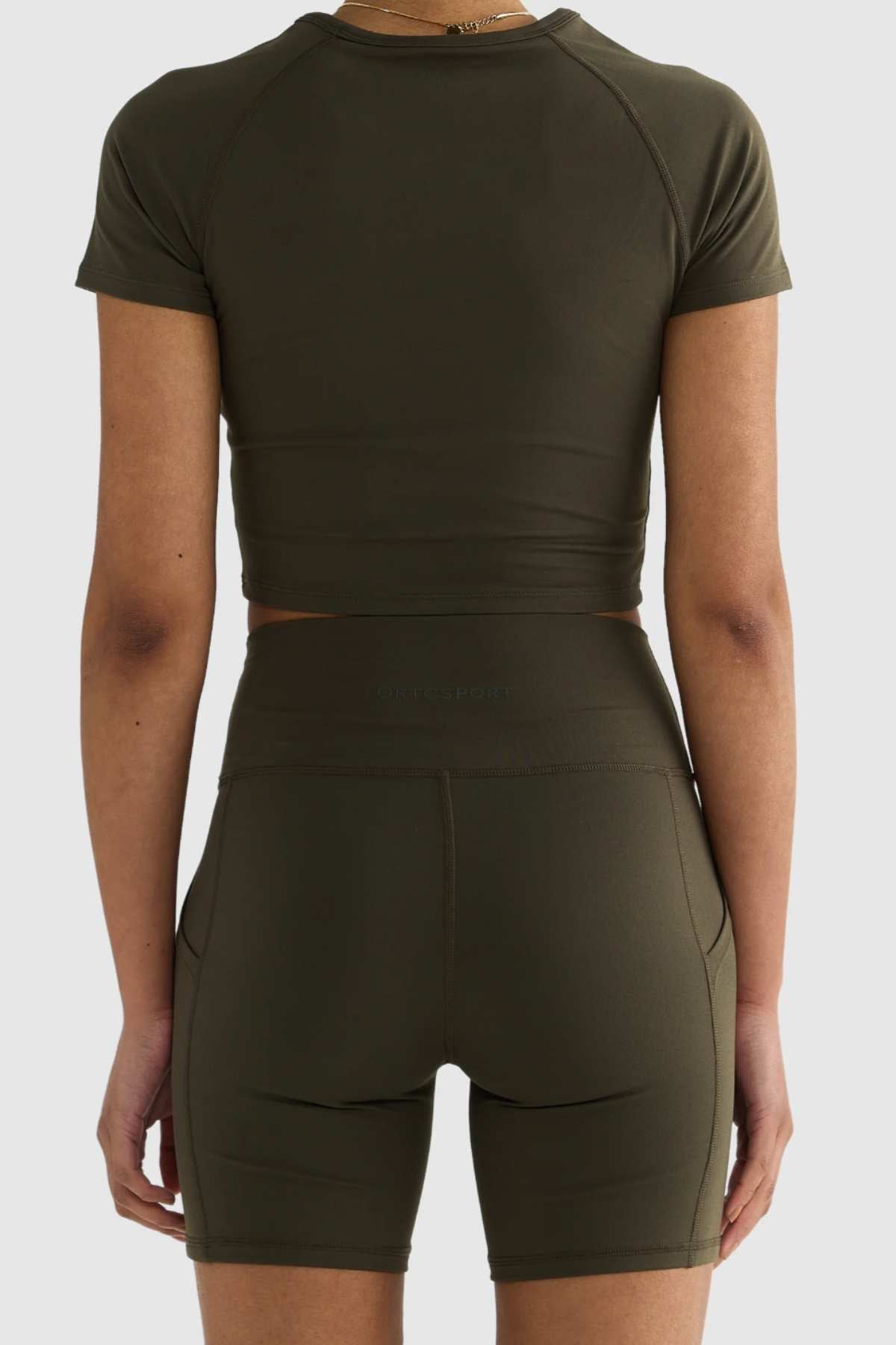 Back view of a person in an olive green short-sleeve crop top paired with matching ORTC Eva 6 Inch Bike Shorts, both made from recycled polyester. The form-fitting outfit stands out beautifully against a plain background.