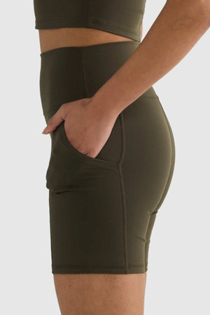 A person dressed in a fitted olive green athletic outfit showcases a side view, with their right hand tucked into the pocket of ORTC Eva 6 Inch Bike Shorts, made from recycled polyester. The high-waisted waistband accentuates the sleek appearance against the plain beige background.