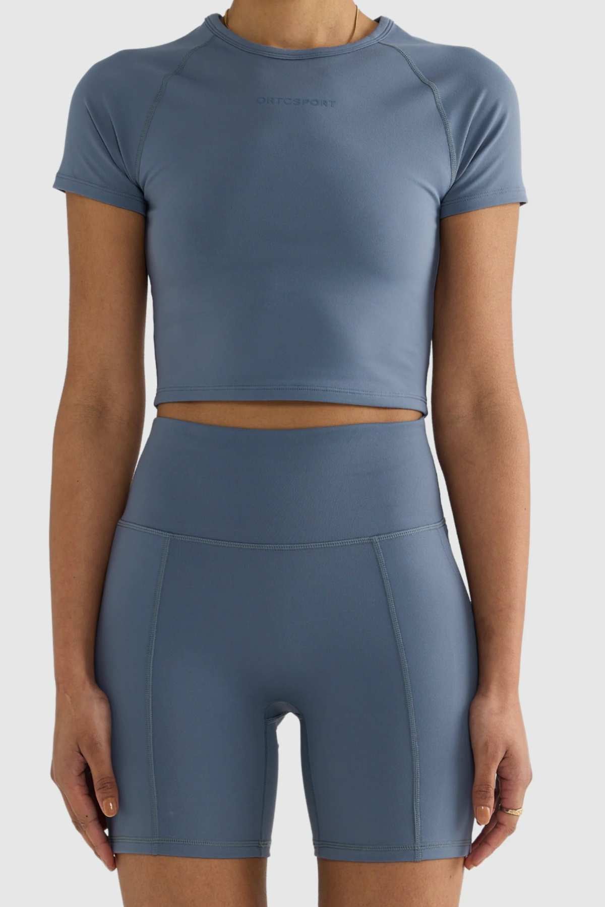 A person stands against a plain background wearing a matching set of blue athletic wear, featuring the ORTC Eva 6 Inch Bike Shorts made from recycled polyester and a short-sleeve crop top. The outfit is form-fitting and minimalist in design.