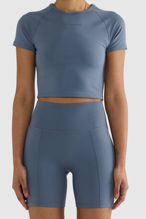 A person stands against a plain background wearing a matching set of blue athletic wear, featuring the ORTC Eva 6 Inch Bike Shorts made from recycled polyester and a short-sleeve crop top. The outfit is form-fitting and minimalist in design.