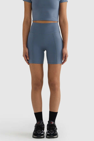 A person wearing a blue crop top and matching ORTC Eva 6 Inch Bike Shorts, crafted from recycled polyester, stands against a plain background. Black socks and black sneakers complete their outfit, with only their body from neck to knees visible.