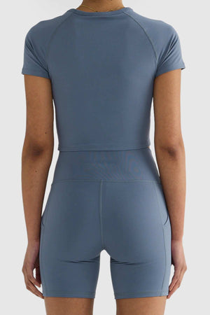 Rear view of a person wearing a matching blue athletic crop top and ORTC Eva 6 Inch Bike Shorts. The clothing is form-fitting and made from smooth, stretchy, recycled polyester fabric. The background is white.