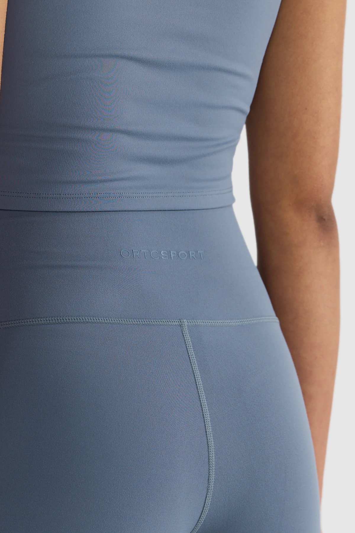 A person in blue athletic wear, featuring ORTC Eva 6 Inch Bike Shorts made from recycled polyester. The ensemble includes a sleeveless top and high-waisted leggings, ideal for sports activities. The material is smooth and stretchy, with the brand name subtly embossed on the waistband.