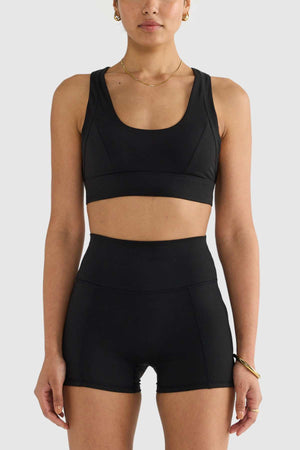 A person wearing a black ORTC Eva Crop Top, designed with brushed fabric, and high-waisted shorts is positioned against a simple light background. Their short hair complements the gold hoop earrings, necklace, and bracelet they are accessorized with, combining style and functionality effortlessly.